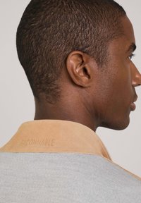 Beige Wildlederkragen auf einem grauen Shirt, das aus strukturiertem Stoff besteht. Der Markenname ist auf der Rückseite des Kragens eingraviert.