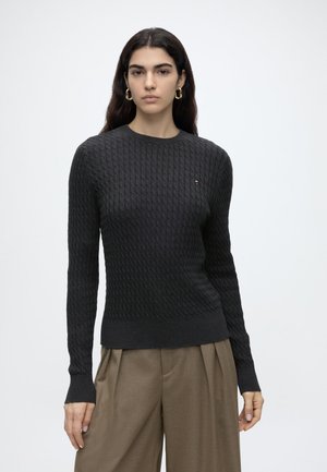 Femme aux longs cheveux foncés porte un pull noir texturé et un pantalon marron plissé sur un fond clair uni.