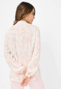 Blusa bianca con motivo floreale rosa, maniche lunghe, polsini elasticizzati e chiusura a bottoni sul retro. Tessuto morbido, vestibilità comoda.