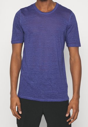 Voksen iført en ensfarvet lilla kortærmet rundhalset T-shirt og sorte bukser, stående med armene afslappet ned langs siderne.