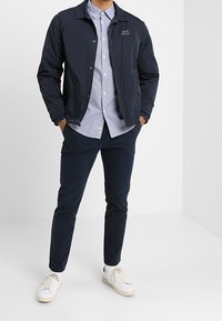 Marineblaue leichte Jacke mit Knopfverschluss und Kragen, darüber ein helles kariertes Hemd. Dunkle Hose und weiße Turnschuhe vervollständigen das Outfit.