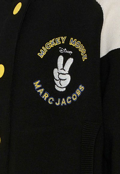 Veste universitaire noire avec un brodé en blanc et jaune représentant Mickey Mouse faisant un signe de paix, avec des boutons jaunes et des poignets côtelés.