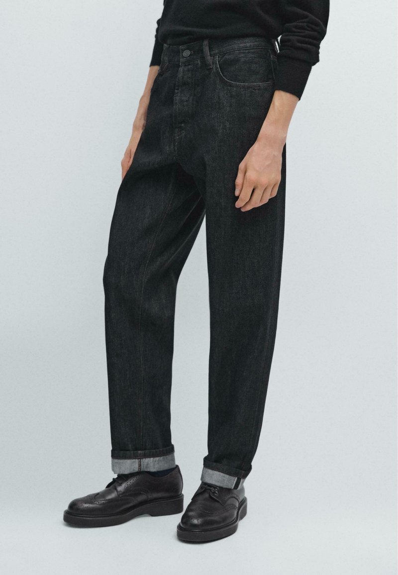 Jeans in denim scuro con una vestibilità ampia, orli risvoltati e accenti di cucitura rossa. Indossati con scarpe in pelle nere e un maglione nero.