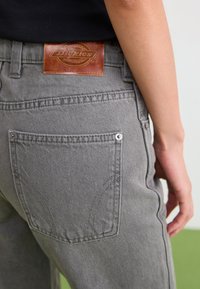 Jeans de mezclilla gris con un parche de marca de cuero en la cintura, bolsillo trasero visible y detalles de costura en contraste.
