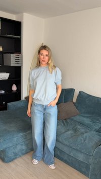 Kvinne med blondt hår i lyseblå collegegenser og vide jeans står ved siden av en turkis sofa med puter i hjørnet av stuen.