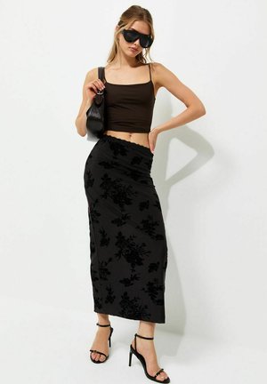 Mujer con top corto negro de tirantes finos, falda midi de terciopelo negro con flores, sandalias negras de tacón alto, gafas de sol y bolso negro.