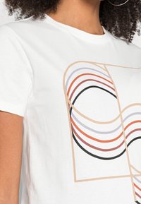 Detail bílého trička s béžově obrysem abstraktních geometrických tvarů a křivkami v černé, oranžové a fialové barvě na hrudi.