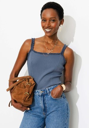 Femme souriante portant un haut bleu sans manches, un jean taille haute, des bijoux en or, tenant un sac à main en daim marron sur fond blanc.