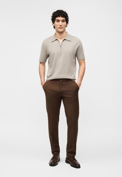 Άνδρας με κατσαρά μαλλιά, όρθιος με τα χέρια στις τσέπες, φορούντας μπεζ πουκάμισο με κοντά μανίκια και γιακά, καφέ παντελόνι και σκούρα καφέ επίσημα παπούτσια.