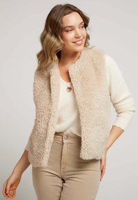 Gilet en fausse fourrure beige avec une texture douce et moelleuse, à l'avant ouvert et col arrondi, porté sur un pull crème et un pantalon taille haute beige.