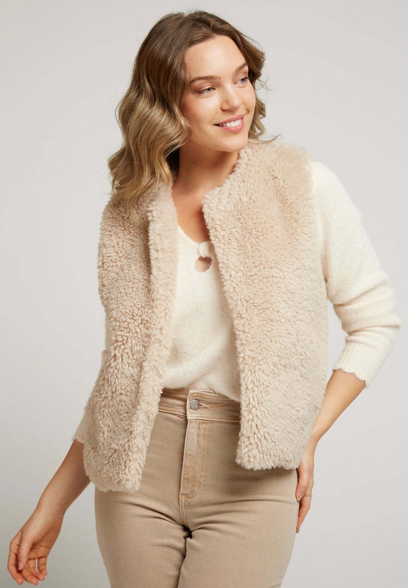Gilet en fausse fourrure beige avec une texture douce et moelleuse, à l'avant ouvert et col arrondi, porté sur un pull crème et un pantalon taille haute beige.
