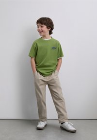 Vihreä lyhythihainen t-paita, jossa valkoinen resorinen pääntie ja sininen graafinen kuvio, yhdistetty beigen värisiin housuihin ja valkoisiin tennareihin.