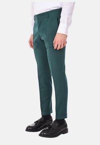 Pantaloni su misura color turchese realizzati in un tessuto liscio, con frontale piatto, gambe con pieghe e orlo arrotolato, abbinati a scarpe nere.