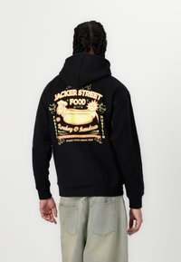 Svart hoodie med en färgglad grafik på baksidan som visar "JACKER STREET FOOD" och en illustration av en korv. Mjuk tyg, avslappnad passform.