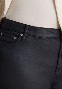 Κοντινό πλάνο της μέσης και της τσέπης μαύρου τζιν, φορεμένο με λευκό ριμπ τοπ και ανοιχτό μπεζ μπουφάν.