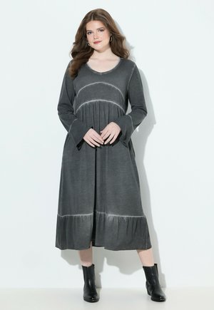 Femme aux longs cheveux ondulés portant une robe midi grise charbon à manches longues et des bottines noires, se tenant contre un fond blanc.