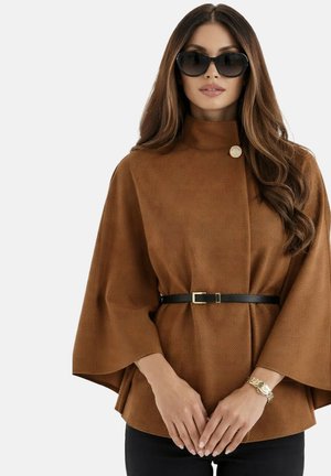 Femme portant une cape marron ceinturée, des lunettes de soleil noires et un bracelet en or, aux cheveux longs et ondulés, sur un fond uni.