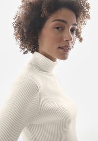 Cremfarbener, gerippter Rollkragenpullover mit taillierter Passform, hohen Kragen und strukturiertem Stoff, gezeigt an einem Modell vor einem schlichten Hintergrund.