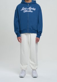 Felpa con zip blu con il testo bianco "Low Lights Studios" sul petto, abbinata a pantaloni della tuta bianchi e sneakers grigie chiare.