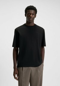 Black short-sleeve t-shirt made of smooth fabric featuring a subtle textured logo on the chest. Paired with light-colored trousers.