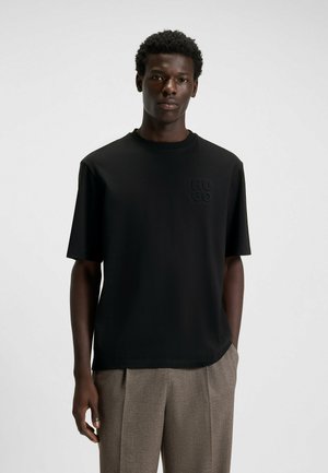 Black short-sleeve t-shirt made of smooth fabric featuring a subtle textured logo on the chest. Paired with light-colored trousers.