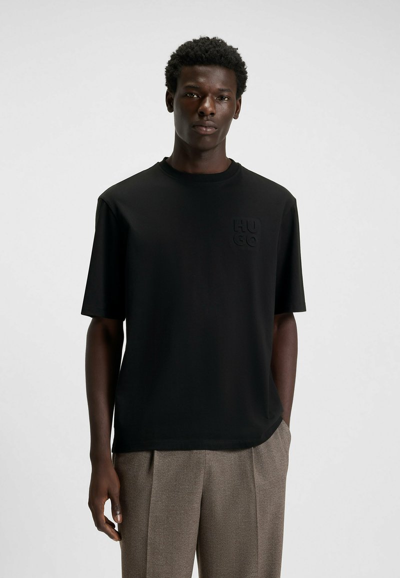 Black short-sleeve t-shirt made of smooth fabric featuring a subtle textured logo on the chest. Paired with light-colored trousers.