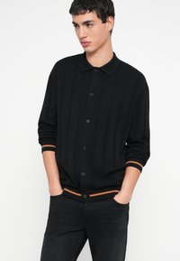 Fekete kötött kardigán bordázott mintával, gombos záródással és galléros nyakkal. Narancssárga díszítésekkel a mandzsettán és az alján.