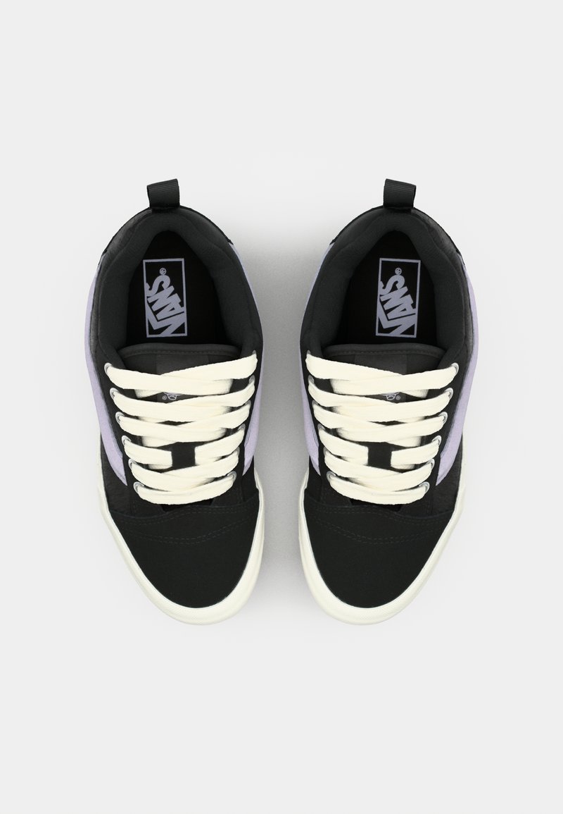 Schwarze und graue Turnschuhe mit lila Akzenten, flachen cremefarbenen Schnürsenkeln und weißen Gummisohlen. Die Zunge zeigt ein Logo und oben ist eine Zuglasche angebracht.