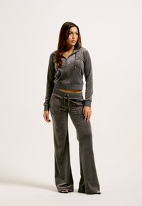 Grijze velours trainingspak met een cropped rits hoodie en wijde broekspijpen. Metallic accenten en trekkoorddetails versterken het ontwerp.