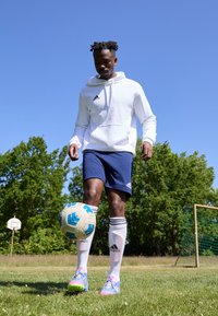 Adidas fehér kapucnis pulóver, tengerészkék rövidnadrág, színes focicipők és fehér zoknik. A játékos ügyesen egy mintás focilabdát egyensúlyoz a füvön.