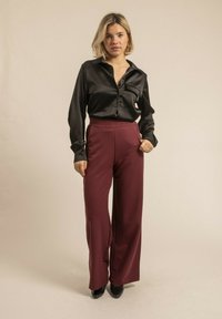 Chemise noire en satin à boutons, à manches longues et avec une poche, associée à un pantalon bordeaux à jambes larges avec un motif à rayures verticales.