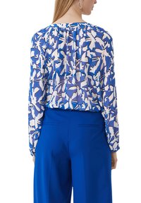 Blaue Blumenbluse mit langen Ärmeln und gerafftem Ausschnitt. Kombiniert mit hochgeschnittenen blauen Hosen, die eine glatte Stoffstruktur zeigen.