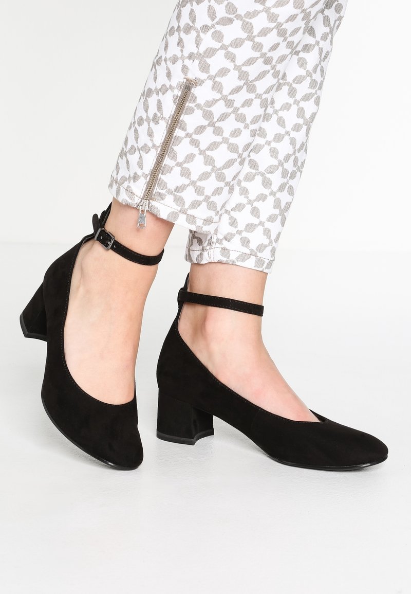 Chaussures basses en daim noir à talon bas avec un bout rond et une bride au niveau de la cheville. Accompagnées de pantalons blancs à motifs avec un ourlet zippé.