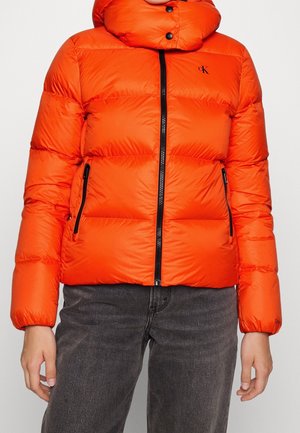 Fel oranje gewatteerde jas met zwarte ritssluiting en zijzakken, gedragen met donkergrijze spijkerbroek, toont bovenlichaam en armen.