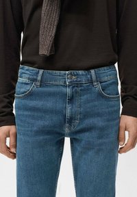 Camiseta de manga larga gris oscuro combinada con jeans de mezclilla azul con diseño clásico de cinco bolsillos, ligero desgaste y cierre frontal de botones.