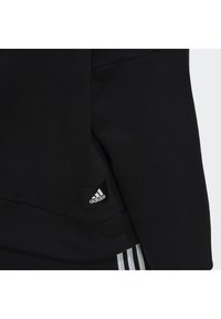 Černá mikina vyrobená z měkké tkaniny, která má malou černou nášivku loga Adidas a bílé akcentační pruhy na spodní části.