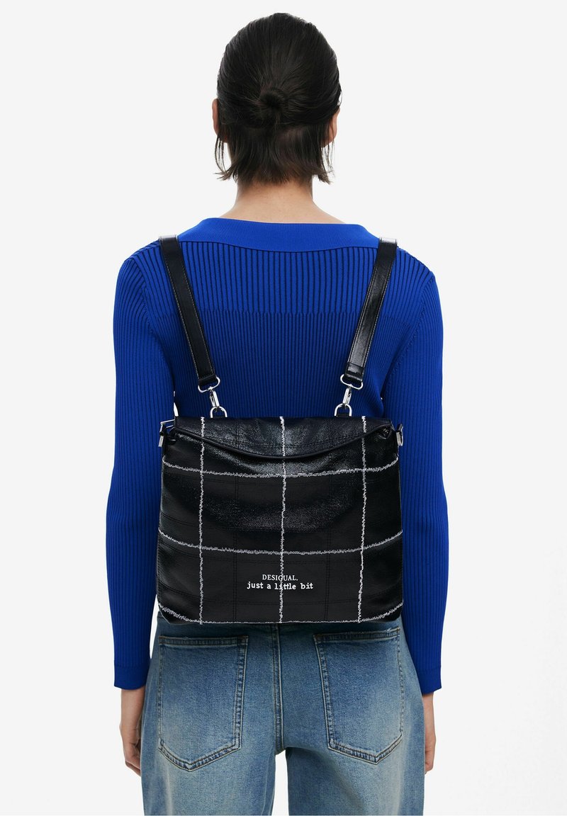 Femme portant un pull côtelé bleu et un jean, portant un sac à dos en cuir noir à carreaux avec une couture blanche et le texte "just a little bit."