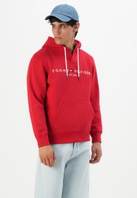 Uomo con felpa rossa Tommy Hilfiger e jeans azzurri, che indossa un cappellino blu, mentre guarda a sinistra su uno sfondo bianco uniforme.