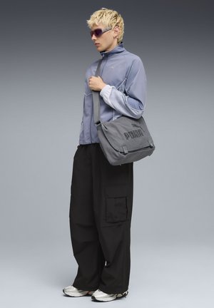 Grijze schoudertas met de tekst "PUMA", gedragen met een lichtblauwe windrunner, zwarte cargobroek en witte sportschoenen. Zilveren zonnebril maakt de look compleet.