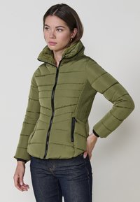 Chaqueta acolchada verde oliva con un cuello alto, costuras horizontales, bolsillos laterales y cremalleras negras. Panel texturizado en el lado izquierdo.