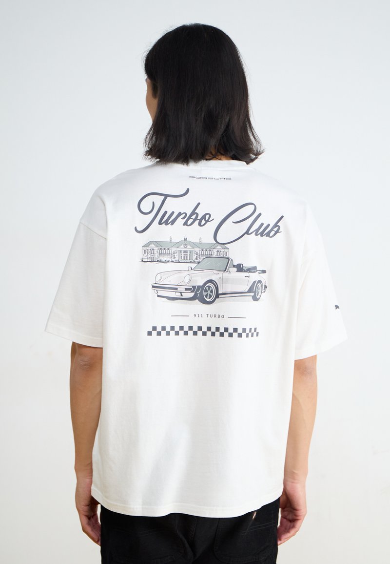 Balts kokvilnas T-krekls ar lielu grafiku aizmugurē, kurā redzams teksts "Turbo Club", Porsche 911 Turbo ilustrācija un šaha klāja rakstu akcenti.