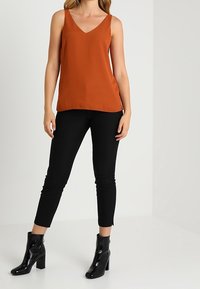 Rostou oranžový bezrukávový top s výstřihem do V a splývavým designem, v kombinaci s černými slim-fit kalhotami a lesklými černými kotníkovými botami.