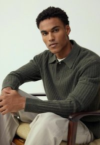 Ciemnozielony, prążkowany sweter z kołnierzykiem i dwoma guzikami. Modelka siedzi w neutralnych, fakturowanych spodniach, z rękami spoczywającymi na kolanach.