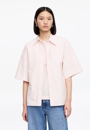 Femme portant une chemise ample à manches courtes rose pâle boutonnée sur un haut blanc et un jean bleu, debout devant un fond blanc.