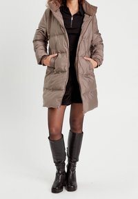 Abrigo acolchado en topo con cremallera frontal, grandes bolsillos y capucha. Llevado sobre un vestido negro y combinado con botas negras hasta la rodilla.