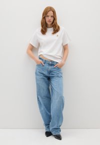 Joven con cabello rojo, vistiendo una camiseta blanca y jeans azules holgados, de pie contra una pared blanca lisa con las manos en los bolsillos.