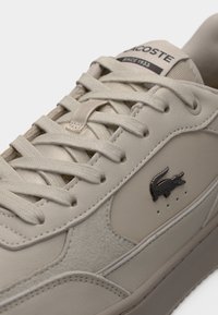 Baskets Lacoste beiges avec lacets texturés, panneaux cousus, logo crocodile en métal et étiquette "Lacoste Depuis 1933" sur la languette.