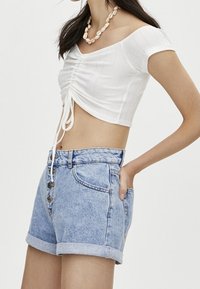 Witte cropped top met geplooide details en koordsluiting, gecombineerd met lichtblauwe high-waisted denim shorts met knoopsluiting en opgerolde zoom.