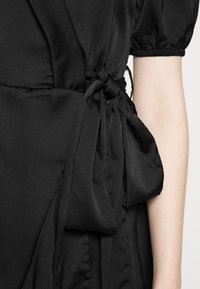 Robe noire présentant une texture douce et lisse avec un détail de taille nouée. Le design comprend des manches bouffantes et une structure de tissu fluide.