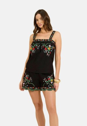 Vrouw draagt een zwart mouwloos topje en korte broek met kleurrijke bloemenborduursel en gegolfde randen, staand met één hand op de heup.
